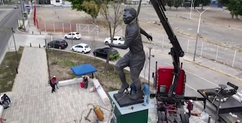 Vista aérea de una estatua de bronce de un hombre con uniforme deportivo siendo levantada por una grúa sobre un pedestal, con trabajadores, vehículos y aceras visibles