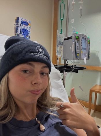 Una mujer joven con gorro y collar en una cama de hospital, muestra el pulgar hacia arriba. Detrás hay un soporte con dos bombas de infusión y bolsas de suero