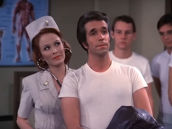 Henry Winkler como Fonzie en una camiseta blanca es examinado por una enfermera con estetoscopio. Hombres en camisetas blancas y un cartel de anatomía detrás