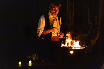 Hombre con barba y cabello gris, camisa blanca y chaleco oscuro, con expresión asombrada, de pie junto a un fuego crepitante en la oscuridad. Dos velas en el suelo