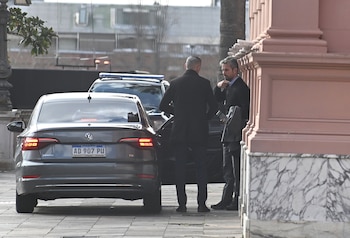 Dos hombres vestidos con traje conversan junto a un vehículo gris con patente argentina en la entrada de un edificio de paredes rosadas