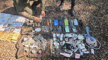 Uma pessoa ajoelhada fiscaliza os itens apreendidos: cigarros, maços de drogas, celulares, carregadores, cabos e armas, sobre um pano plano sobre chão arenoso.