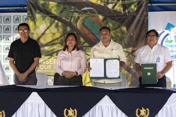 Cuatro personas, tres hombres y una mujer, de pie detrás de una mesa. Dos hombres sostienen documentos y carpetas. Hay banners con logotipos de CONAP y un quetzal