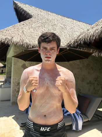 Joven con protector solar blanco en cara y torso, vestido con bañador negro, haciendo puños, frente a un edificio con techo de paja y sombrilla