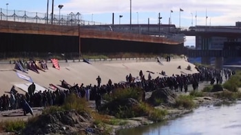 Gran grupo de personas junto a un río y un terraplén de hormigón inclinado, con un paso elevado y una valla con alambre de púas en segundo plano