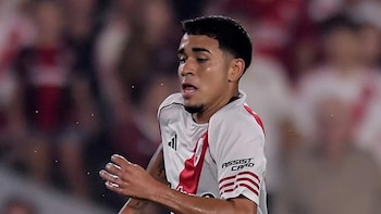 Vista frontal de un jugador de fútbol con camiseta blanca y roja número 19, driblando el balón con dos jugadores de camiseta verde a su alrededor en un campo de césped