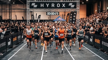 Varios corredores con dorsales avanzan bajo una estructura metálica con la marca Hyrox y un cronómetro digital rodeados por público.