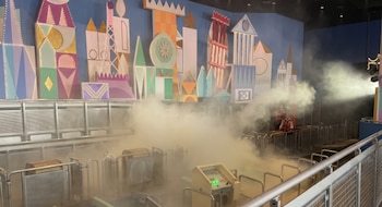 Humo blanco cubriendo la zona de embarque de una atracción. Al fondo, una pared azul con figuras geométricas coloridas de edificios. Barandas metálicas y una consola de control
