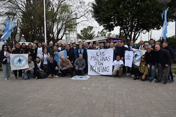 Grupo de personas en una plaza posando junto a una pancarta blanca que dice Las Malvinas son argentinas y varias banderas de Argentina