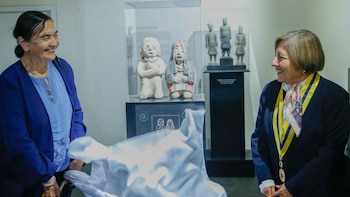 Dos mujeres retiran una tela blanca frente a una vitrina con dos estatuillas antropomorfas en una sala de museo
