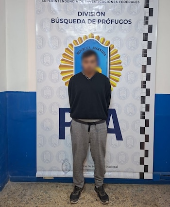 Hombre con el rostro borroso viste suéter negro y pants grises, de pie frente a un cartel oficial de la División Búsqueda de Prófugos de Argentina con una escala de altura