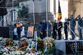 Oficiales de policía uniformados realizan el saludo militar frente a un retrato y arreglos florales junto a una bandera belga