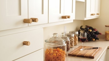 Muebles blancos de cocina con corchos de vino como tiradores, frascos con alimentos, una copa y una tabla de madera sobre la encimera.