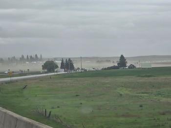 Paisaje rural con campos de hierba, carretera con vehículos y árboles bajo una densa capa de polvo o niebla. Cielo nublado