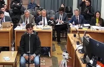 Un hombre sentado frente a un micrófono en una sala judicial con personas de traje en escritorios y policías de pie detrás