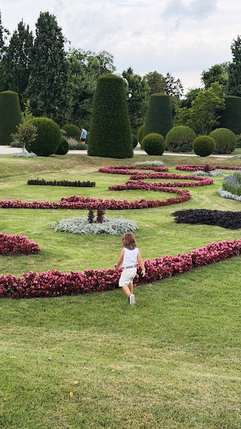 Niño de espaldas en un jardín con césped, sendas de flores rosadas, arbustos con formas y árboles altos bajo un cielo nublado. Otra persona al fondo
