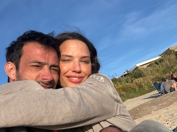 Una pareja sonriente se abraza en primer plano, con el cielo azul, vegetación y edificios a lo lejos, y otras personas en la arena