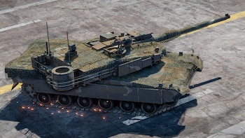 Tanque militar con camuflaje de red y pintura, cañón largo, orugas, antenas y una ametralladora, sobre hormigón con puntos de luz