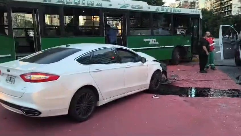 Un Ford Fusion blanco con el frontal dañado tras colisionar con un autobús verde de la línea 70, mientras dos hombres observan la escena y líquidos se esparcen por la calzada.