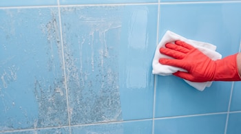 Una mano con un guante rojo sostiene un paño blanco sobre azulejos azules que presentan manchas de sarro a la izquierda.
