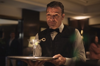 Hombre vestido con traje de mayordomo y moño, sostiene una bandeja plateada con una copa de martini con aceituna