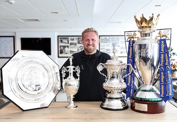 Kasper Schmeichel sonríe detrás de una mesa de madera con cuatro trofeos de fútbol, entre ellos la copa de la Premier League