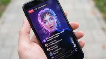 Mano sostiene un teléfono celular mostrando una transmisión en vivo con una influencer virtual, número de espectadores, comentarios y corazones flotantes.