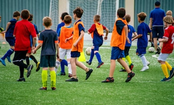 Grupo de niños en camisetas, pantalones cortos y chalecos deportivos sobre un campo de césped artificial con un balón de fútbol y una portería