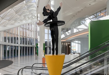 Escultura negra estilizada de una figura en equilibrio sobre un pedestal amarillo, con brazos alzados, en el atrio de un edificio moderno con techos de cristal y escaleras mecánicas