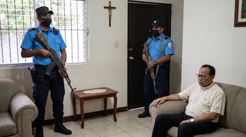 Dos policías uniformados con gorras, mascarillas y fusiles de pie en una sala. Un sacerdote con camisa clerical y gafas está sentado en un sofá.