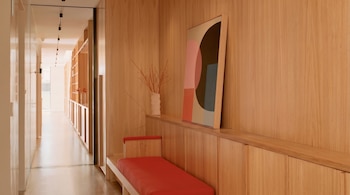 Un pasillo interior con paredes y estanterías de madera clara, un banco con cojines rojos, una pintura abstracta y un jarrón decorativo sobre una repisa.