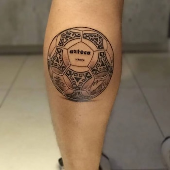 Pierna humana con un tatuaje de un balón de fútbol blanco y negro. El balón presenta patrones geométricos y el texto 'azteca' y 'México'