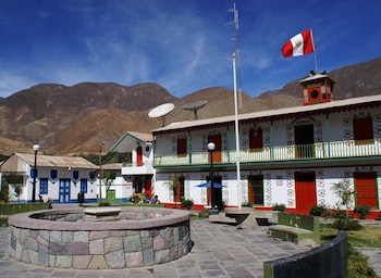 Una plaza de piedra con una fuente circular rodeada de edificios blancos pintados con motivos de colores y la bandera de Perú