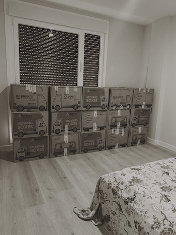 Cajas de cartón apiladas, una ventana con persiana bajada, paredes lisas, suelo de madera claro y parte de una cama con edredón floral