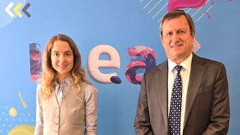 Una mujer con cabello rizado castaño claro y un hombre de traje gris con corbata a lunares posan sonriendo frente a una pared azul con el logo de IDEA