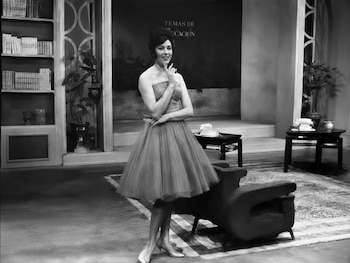 Fotografía en blanco y negro de Julissa de pie en un estudio, vistiendo un vestido strapless de falda voluminosa y tacones, con estanterías y muebles al fondo