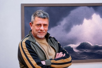 Un hombre con chaqueta deportiva y brazos cruzados posa delante de una pintura enmarcada de un mar embravecido.