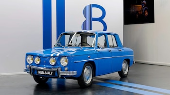 Un automóvil clásico azul con dos franjas blancas y cuatro faros delanteros, estacionado frente a un mural con el número ocho