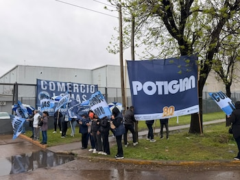 Personas con banderas celestes y blancas junto a carteles con los nombres Potigian y Comercio Lomas frente a un depósito industrial