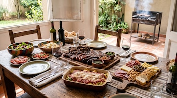 Mesa de madera con carnes a la parrilla, pastas, embutidos, ensaladas, copas de vino tinto y vajilla junto a un patio con parrilla.