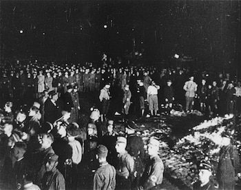 Imagen nocturna en blanco y negro de una gran multitud observando una pira de libros ardiendo en el suelo, con varias personas llevando uniformes militares
