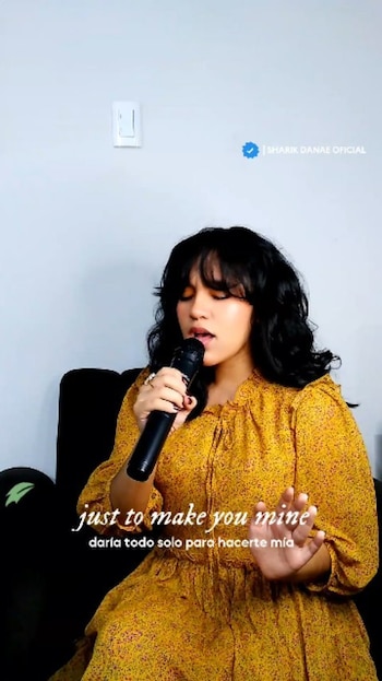 Joven mujer con cabello oscuro y flequillo, vestida de amarillo estampado, canta en un micrófono negro, su mano derecha levantada