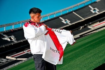 Hombre joven con chaqueta blanca, roja y negra sostiene camiseta de fútbol blanca con franja roja, nombre "ALMADA" y "DIRECTV". Campo verde, gradas negras y cielo azul de fondo