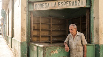 Un hombre mayor junto al mostrador de madera de una tienda con estantes vacíos y un letrero con la frase Bodega La Esperanza.