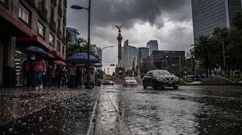 Calle urbana con agua y granizo en el suelo, coches circulando, peatones con paraguas, edificios y el monumento del Ángel de la Independencia bajo un cielo oscuro.