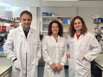 Tres profesionales con batas blancas de laboratorio posan sonriendo. Un hombre está a la izquierda y dos mujeres a su derecha, en un laboratorio con estantes y equipos