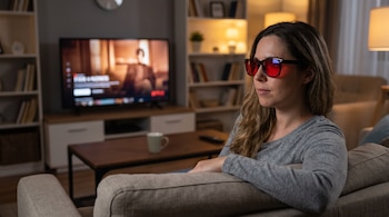 Mujer sentada en un sofá gris con gafas de cristales rojos, mirando una televisión encendida que muestra una serie, en una sala de estar.