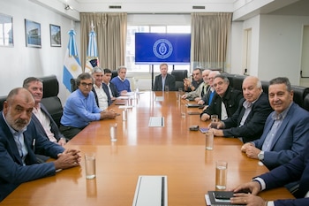 Un grupo de trece hombres en un despacho, sentados alrededor de una mesa de madera. Al fondo, dos banderas argentinas y una pantalla con el logo del Ministerio de Economía