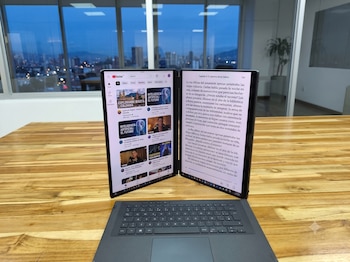Portátil ASUS ROG Zephyrus Duo con dos pantallas abiertas, una muestra YouTube y la otra un e-book, sobre una mesa de madera, con una ciudad de noche al fondo