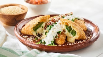 Primer plano de quesadillas fritas infladas, partidas por la mitad con queso fundido y hoja santa, servidas en un plato de barro con guarniciones.